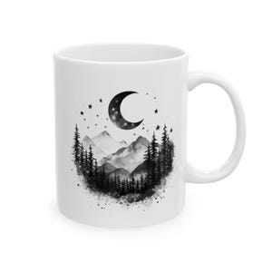 Może przedstawiać: Biały ceramiczny kubek z czarno-białą ilustracją księżyca w kształcie półksiężyca, gwiazd, gór i sosen. Projekt ma efekt akwareli, tworząc marzycielski, niebiański motyw. Kubek ma standardowy uchwyt.