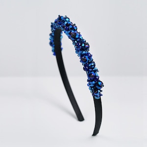 May include: A black headband decorated with bright blue beads. The beads are clustered, forming a textured, striking design. The headband is set against a plain white background, highlighting the accessory's details.