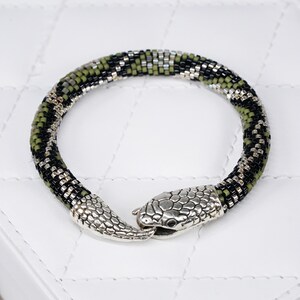 May include: A snake-shaped bracelet made from green, black, and silver beads. The snake's head and tail are silver-coloured metal, featuring detailed scale patterns. The bracelet is designed to wrap around the wrist.