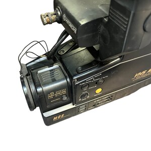 May include: Black Hitachi camcorder with a silver MOS logo and a black lens with the text "X6 ZOOM VIDEO ZOOM LENS 1:1.6 12~72 mm MACRO" printed on it. The camcorder has a variety of buttons and controls, including power, focus, iris, date, and self timer.