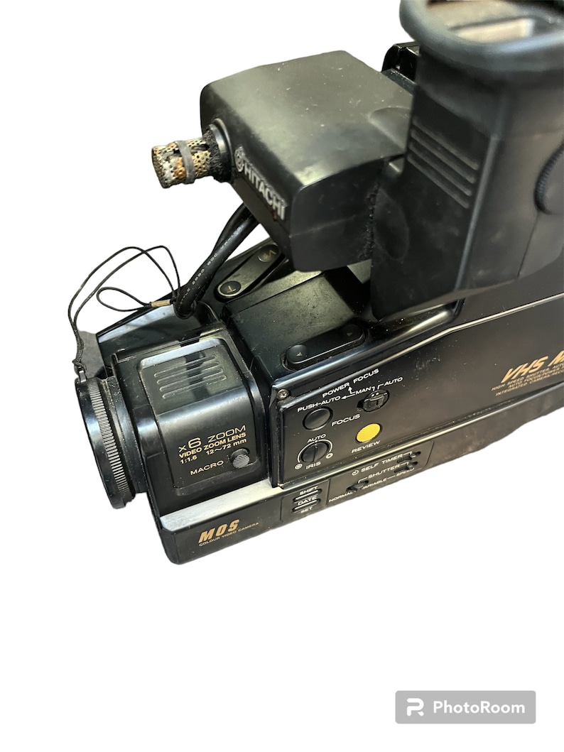 May include: Black Hitachi camcorder with a silver MOS logo and a black lens with the text "X6 ZOOM VIDEO ZOOM LENS 1:1.6 12~72 mm MACRO" printed on it. The camcorder has a variety of buttons and controls, including power, focus, iris, date, and self timer.