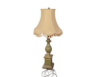 May include: A green and gold table lamp with a cream-colored lampshade with a fringe trim. The lamp has a decorative base and a cord with a plug.