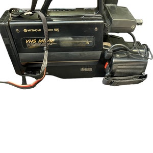 May include: A black Hitachi VHS Movie camcorder with a built-in microphone and a black strap. The camcorder has a silver trim and the text "VHS Movie" and "X6 Zoom" printed on it.
