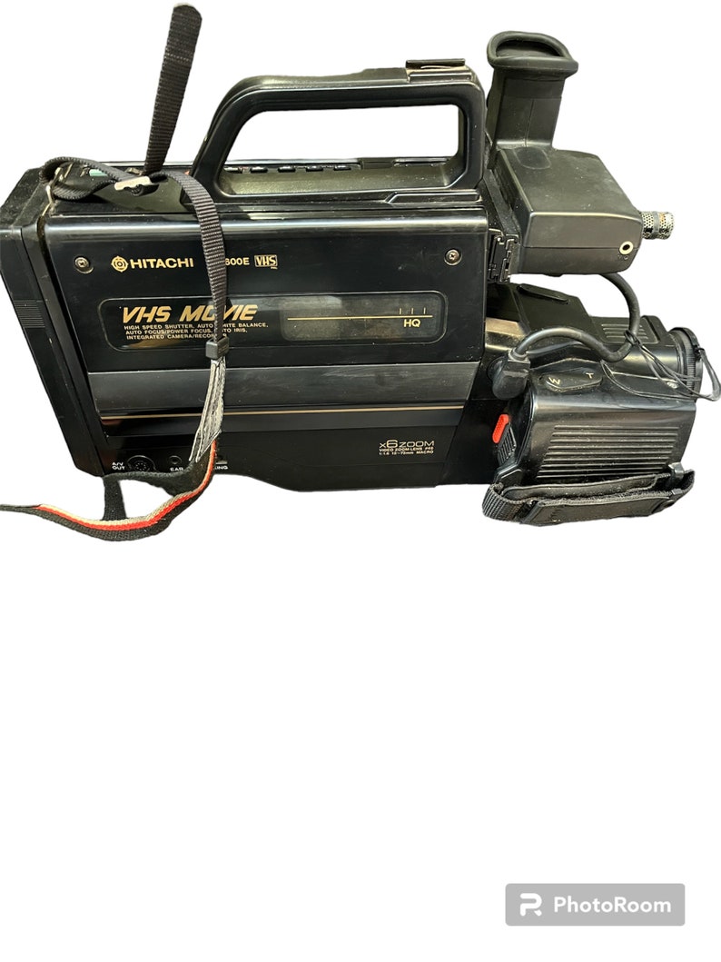 May include: A black Hitachi VHS Movie camcorder with a built-in microphone and a black strap. The camcorder has a silver trim and the text "VHS Movie" and "X6 Zoom" printed on it.