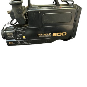 May include: Black VHS Movie 600 camcorder with a built-in camera recorder. The camcorder has a zoom lens, a viewfinder, and a handle.