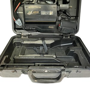 May include: A black VHS camcorder with a 16x zoom lens in a black hard case with a handle. The camcorder has a red and white strap and the text "VHS MOVIE" on the front.
