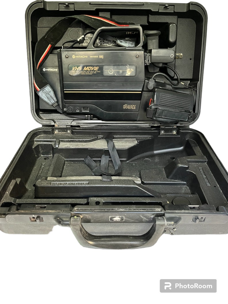 May include: A black VHS camcorder with a 16x zoom lens in a black hard case with a handle. The camcorder has a red and white strap and the text "VHS MOVIE" on the front.
