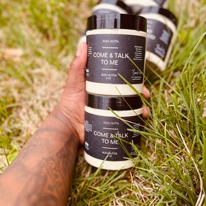 May include: Three jars of body butter labeled "Soul Glitta Come & Talk To Me" with black text on a white background. The jars are stacked on top of each other and are sitting on a bed of green grass.