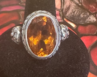 May include: Silver ring featuring an oval, amber-colored gemstone. The ring has a detailed silver band with decorative elements on each side of the gemstone. The gemstone is faceted, catching the light and displaying a warm, golden hue.