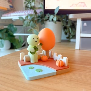 May include: A small figurine set featuring a pale green character playing a dark green guitar while sitting on a log. The scene includes a large orange tree, two small orange pumpkins, a white picket fence, and a light blue pond with lily pads.