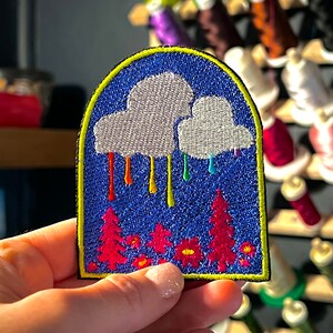以下が含まれることがあります： 虹色の雨粒がピンクの樹木の森に降っている白い雲が描かれたカラフルな刺繍パッチ。パッチは黄色の縁取りで囲まれており、青い背景になっています