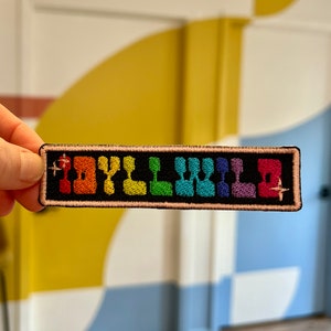 May include: A rainbow-colored embroidered patch with the word "HOLLYWOOD" in a pixelated font. The patch is on a black background and has a pink border.