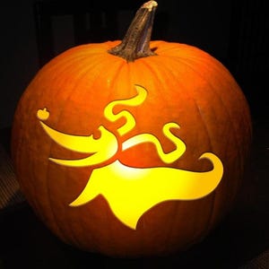 May include: A round orange pumpkin with a carved design of a ghost dog featuring a long snout, floppy ears, and a flowing body. The interior is illuminated, causing the cutout shape to glow with a bright yellow light against the dark rind.