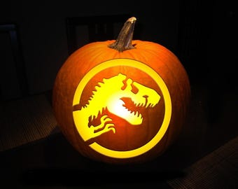 May include: A carved orange pumpkin featuring a glowing yellow silhouette of a Tyrannosaurus Rex dinosaur inside a circular border. The design is illuminated from within, creating a bright contrast against the dark orange pumpkin skin.