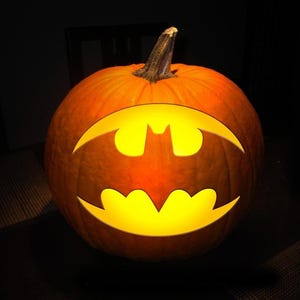 May include: A carved orange pumpkin with a glowing Batman logo. The pumpkin is lit from within, creating a bright yellow silhouette of the iconic bat symbol. The background is dark, emphasizing the illuminated pumpkin.