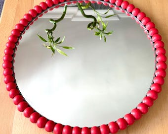 May include: A circular mirror featuring a decorative frame composed of a single row of smooth, matte red wooden beads. The reflective surface of the mirror shows a clear reflection of a green plant with thin stalks, capturing the light and surroundings.