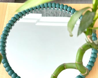 May include: A circular mirror featuring a frame composed of numerous small, matte teal-colored wooden beads arranged in a continuous ring around the reflective glass surface, creating a textured border for the round mirror design.