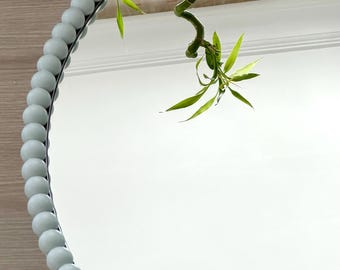 May include: A round wall mirror featuring a frame composed of smooth, light mint green spherical beads. The reflective surface shows a clear view of a green bamboo plant with slender leaves, reflecting the plant's stalk and foliage in the glass.