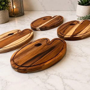 May include: Four oval-shaped wooden serving boards with a natural brown wood grain finish. Each board features a distinct thumb hole cutout on one side, resembling an artist's palette, and a carved groove running along the perimeter of the surface.