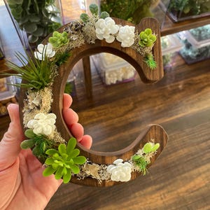 May include: A wooden letter "C" decorated with white and green faux succulents, moss, and small white flowers. The letter is held in a hand, with a dark wood background.