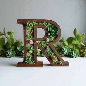 May include: A wooden letter "R" is filled with green moss and small succulents. The letter is a dark brown color and has a natural wood grain. The succulents are various shades of green and pink, adding a touch of color to the design. The letter is a decorative item.