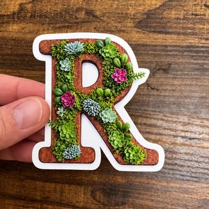 Puede incluir: Una pegatina troquelada con la forma de la letra R, que presenta un fondo con textura de veta de madera marrón decorado con varias ilustraciones en miniatura de musgo verde y plantas suculentas en tonos azul claro, verde lima y rosa brillante.