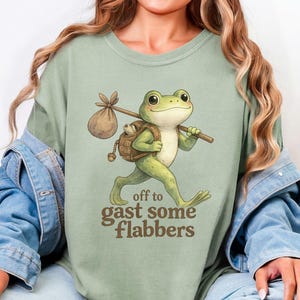 May include: Sage green t-shirt featuring a cartoon frog with a backpack and walking stick. The frog is walking with the text "off to gast some flabbers" below. The shirt is made of a soft material.