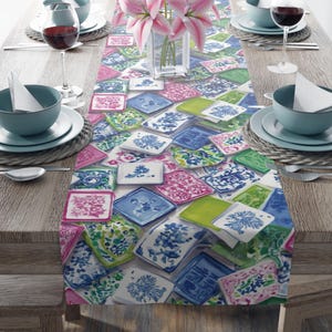 May include: A rectangular table runner featuring a repeating pattern of square tiles in shades of blue, pink, and green, decorated with floral and geometric motifs. The runner is placed on a wooden table set with blue bowls, wine glasses, and flowers.