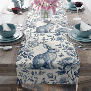 May include: A blue and white table runner with a floral and bunny pattern. The runner is on a wooden table with blue and white dishes and glasses.