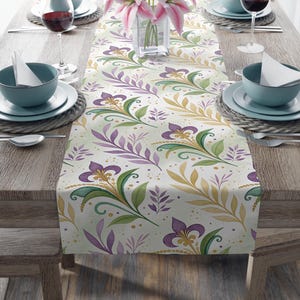 May include: A floral table runner with a watercolor design featuring purple, green, and gold botanical elements. The runner is placed on a wooden table set for a meal, with plates, bowls, and wine glasses.