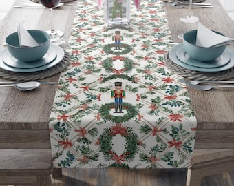 May include: A festive table setting featuring a white table runner with a repeating pattern of green wreaths, red bows, and nutcrackers. The table is set with teal-colored bowls, plates, and wine glasses, creating a holiday theme.