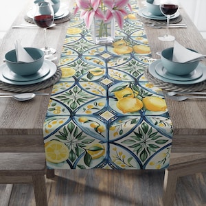 May include: A white table runner with a blue and green geometric pattern featuring lemons and leaves. The runner is on a wooden table with blue bowls and wine glasses.