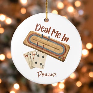 May include: A round white ceramic ornament featuring a wooden cribbage board with blue and red tracks, four pegs, and a hand of playing cards. The text "Deal Me In" is printed in brown above the board, and the name "PHILLIP" is printed below.
