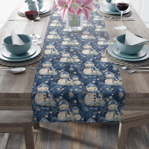 May include: A blue table runner with a repeating pattern of snowmen wearing blue hats and scarves. The snowmen have button eyes and twig arms. The runner is on a wooden table set for a meal, with plates, bowls, and wine glasses.