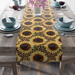 May include: A table runner featuring a sunflower and leopard print design. The runner displays large yellow sunflowers with brown centers against a brown leopard print background. The runner is placed on a wooden table, with place settings and a vase of pink flowers.