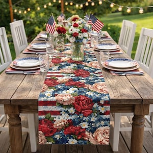 May include: A wooden table set for a patriotic celebration, featuring a floral table runner with red, white, and blue flowers and stripes. American flags and flower arrangements adorn the table, with white chairs and string lights in the background.