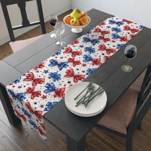 May include: A white table runner with a pattern of red and blue bows and stars. The runner is placed on a dark wooden table, with a plate, silverware, and wine glasses visible.