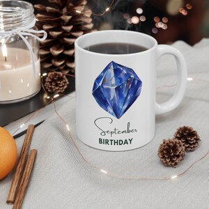 May include: White ceramic mug with a blue gemstone illustration and the text "September BIRTHDAY" in black and green lettering.