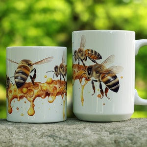 May include: Two white ceramic mugs with a design of bees and honeycombs. The larger mug has a handle and the smaller mug does not.