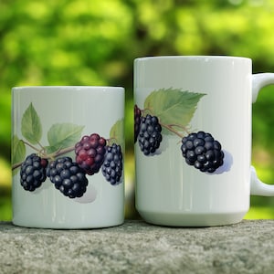 May include: Two white ceramic mugs with blackberries and green leaves printed on them. The mugs are sitting on a gray stone surface.