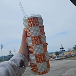 May include: A white and orange checkered tumbler with rhinestones. The tumbler has a straw and is being held by a hand.