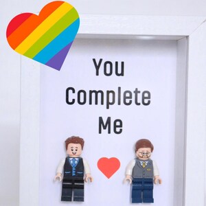 Könnte beinhalten: Ein weißer Rahmen mit einem Regenbogenherz oben links. Im Rahmen befinden sich zwei Lego-Figuren, ein rotes Herz und der Text "You Complete Me" und "Hannah & James".
