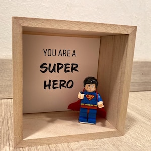 Könnte beinhalten: Eine Holzkiste mit einer weißen Karte im Inneren, auf der "You are a super hero" steht. Eine kleine Superman Lego-Figur steht vor der Karte.