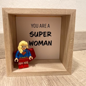 Könnte beinhalten: Ein Holzrahmen mit einer Karte mit der Aufschrift "YOU ARE A SUPER WOMAN". Eine Lego-Figur einer Superheldin mit blonden Haaren, einem blauen Oberteil, einem roten Rock und einem roten Umhang befindet sich vor der Karte.