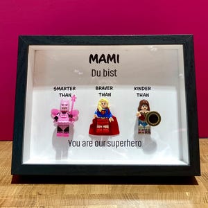 Könnte beinhalten: Ein gerahmtes Bild mit dem Text "Mami Du bist" und drei Lego-Minifiguren. Der Text unter den Figuren lautet "Kluger als, Mutiger als, Freundlicher als, Du bist unser Superheld".