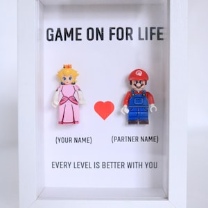 Könnte beinhalten: Ein weißer Bilderrahmen mit einer klaren Glasscheibe. Im Rahmen befinden sich zwei Lego-Minifiguren, eine von Prinzessin Peach und eine von Mario. Der Rahmen trägt den Text "GAME ON FOR LIFE" oben und den Text "(YOUR NAME)" und "(PARTNER NAME)" unter den Figuren. Der Text "EVERY LEVEL IS BETTER WITH YOU" befindet sich unten am Rahmen.