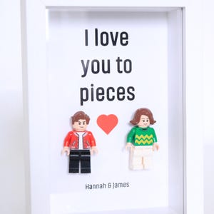 Könnte beinhalten: Ein gerahmtes Kunstwerk mit dem Text "I love you to pieces" über zwei Lego-Figuren und einem roten Herzen. Die Lego-Figuren tragen eine rote Jacke und einen grünen Pullover. Die Namen "Hannah & James" stehen darunter.