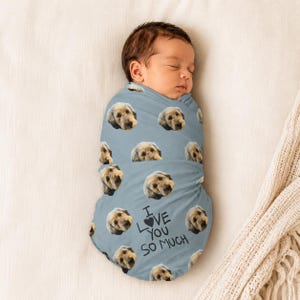 May include: A baby wrapped in a pale blue swaddling blanket featuring a repeating pattern of a dog's face. The blanket has the handwritten text "I love you so much" in black. The baby is asleep.