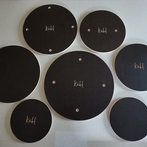 Puede incluir: Un conjunto de siete posavasos circulares negros con bordes blancos. Cada posavasos tiene un remache metálico y el texto manuscrito "k&H". Los posavasos varían en tamaño, el más grande mide aproximadamente 20 cm de diámetro.