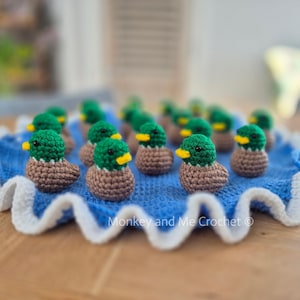 May include: A group of miniature crocheted ducklings, each with a green head, brown body, and yellow beak. They are displayed on a blue crocheted mat with a white, frilled border. The image highlights the handmade quality of the items.